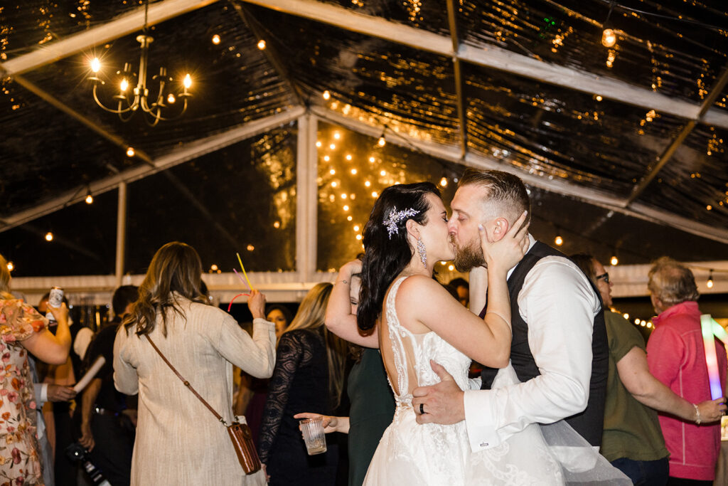 A couple kisses on the dance floor under a tent adorned with fairy lights, as guests around them capture the moment and enjoy the celebration.