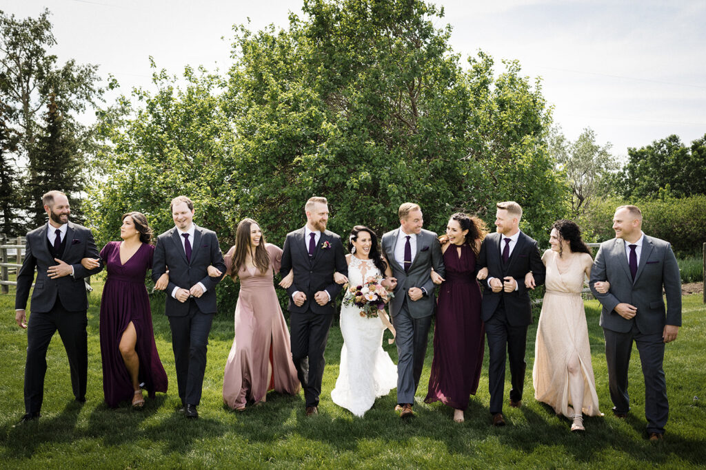 A wedding party posing outdoors, with couples in formal attire smiling and holding hands.