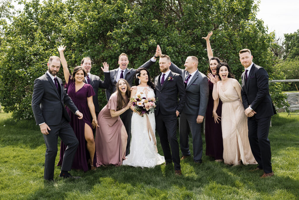 A wedding party posing joyfully on a grassy lawn with some members raising their arms and making playful expressions.