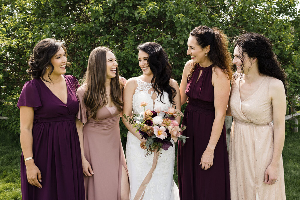A bride and her bridesmaids sharing a cheerful moment outdoors, with the bridesmaids dressed in coordinating dresses while the bride holds a bouquet.