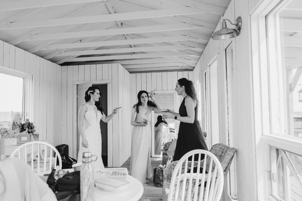 Three women having a conversation in a white wooden room, two appear to be bridesmaids while one is wearing a wedding dress.