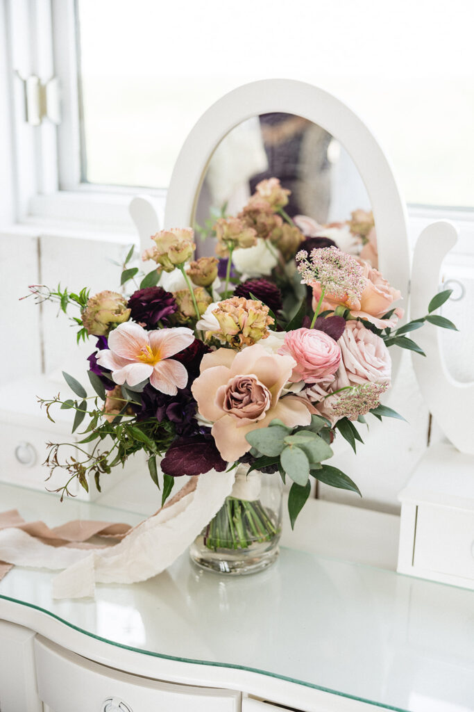 A bouquet of assorted flowers in a glass vase placed on a white vanity table with an oval mirror. A Garden Wedding with Pops of Plum