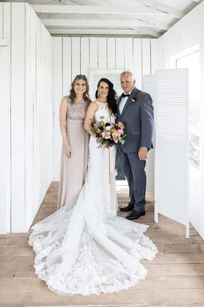 A bride flanked by her smiling parents inside a room with white wooden walls.