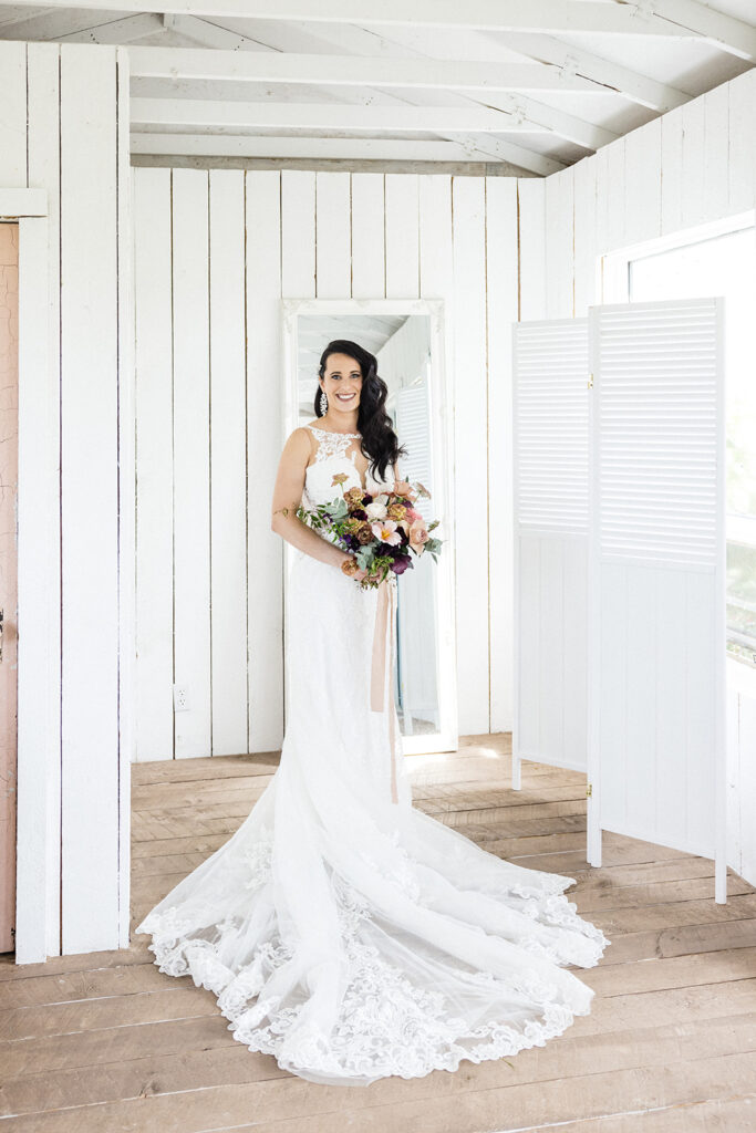 A bride in a white gown and veil, holding a bouquet, smiling in a room with white walls and wooden flooring.