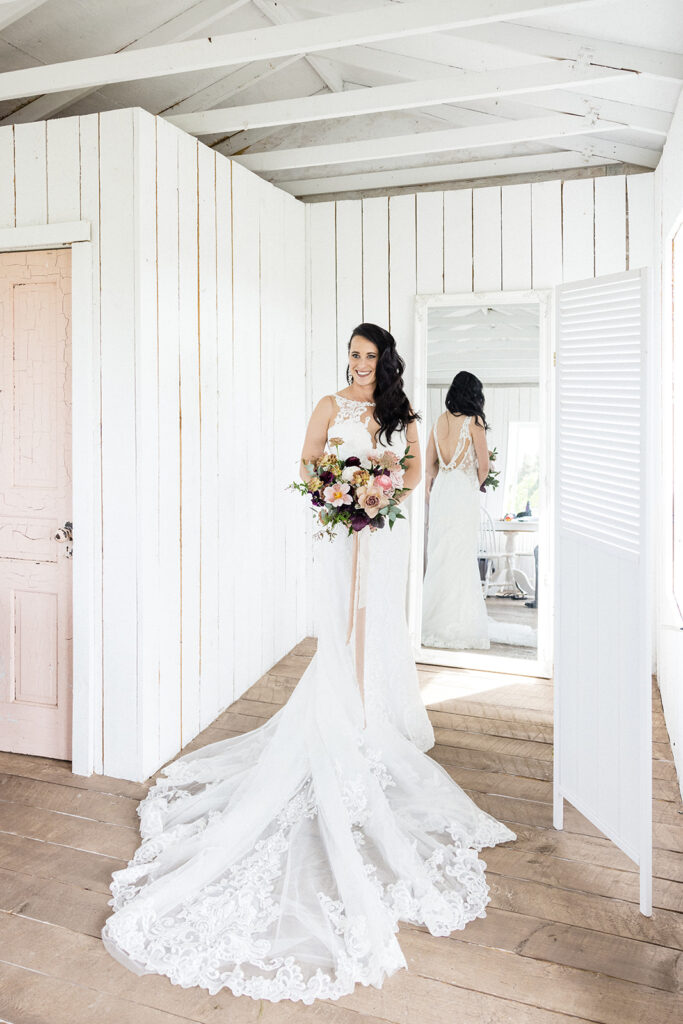 A bride in a white dress holding a bouquet of flowers, standing in a room with rustic white wooden walls and a mirror reflecting her image.