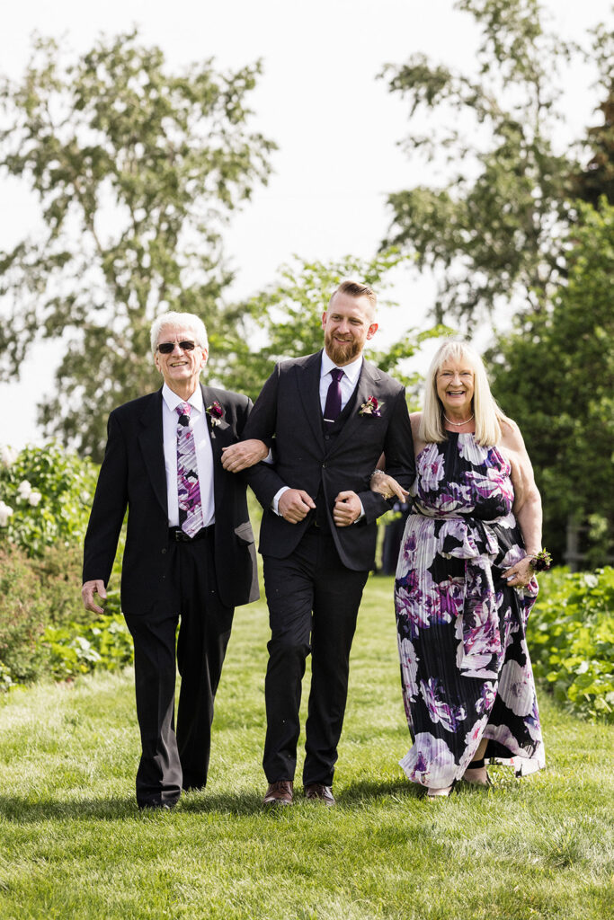 A groom walks arm in arm with his parents on a grassy path during a sunny wedding day.
