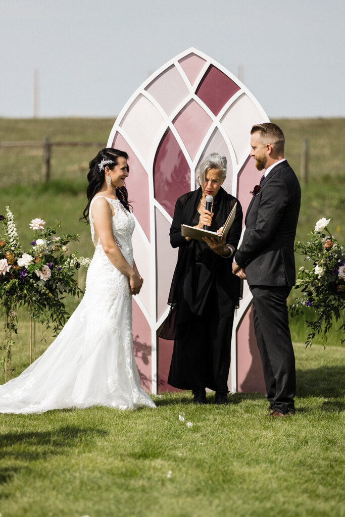 A wedding ceremony taking place outdoors with a bride and groom exchanging vows in front of an officiant, with a decorative arch and floral arrangements in the background. A Garden Wedding with Pops of Plum