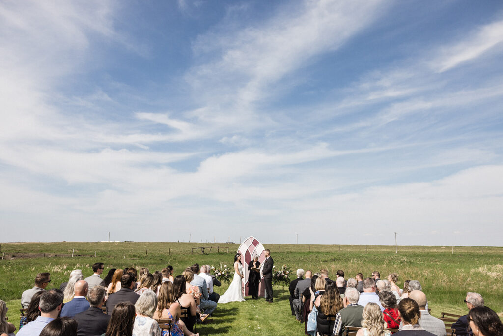 Outdoor wedding ceremony with guests seated on chairs facing a couple standing under an arch on a sunny day with open fields in the background.