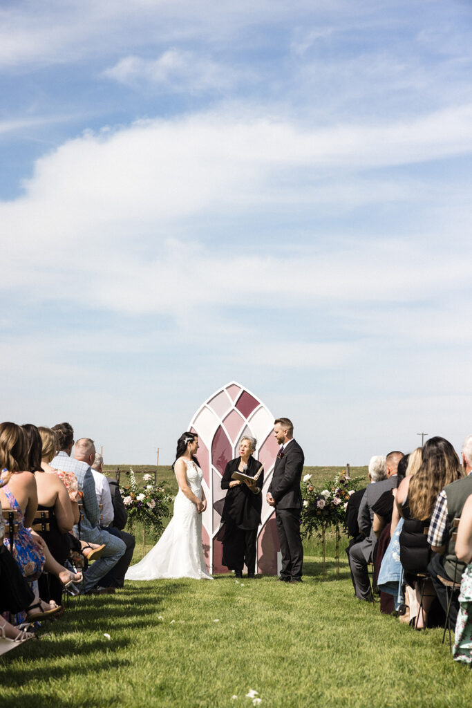 A bride and groom stand facing each other during an outdoor wedding ceremony with guests seated on either side.