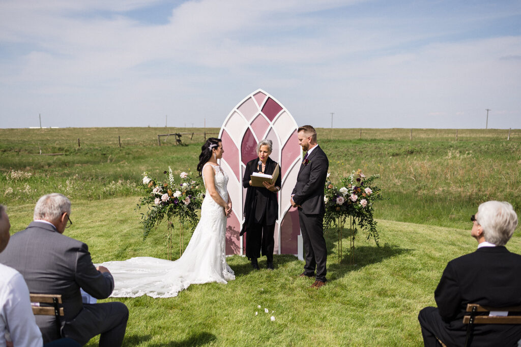A wedding ceremony taking place outdoors with the bride and groom standing in front of an officiant, with guests seated on either side.