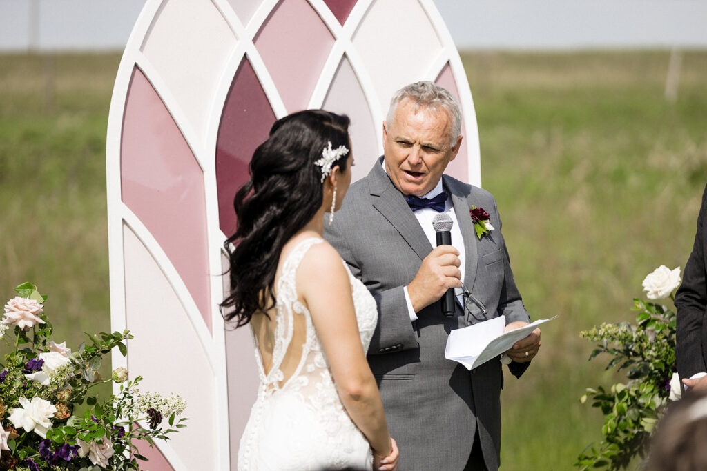 A bride listening to a man giving a speech at an outdoor wedding ceremony.