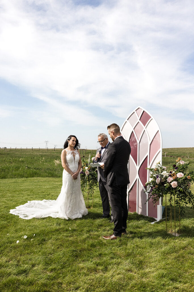 Bride and groom stand before an officiant at an outdoor wedding ceremony. A Garden Wedding with Pops of Plum