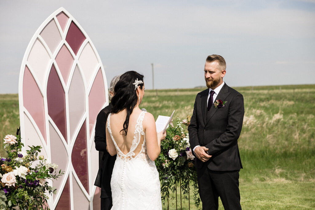 A bride and groom exchange vows outdoors with a decorative backdrop and floral arrangements.