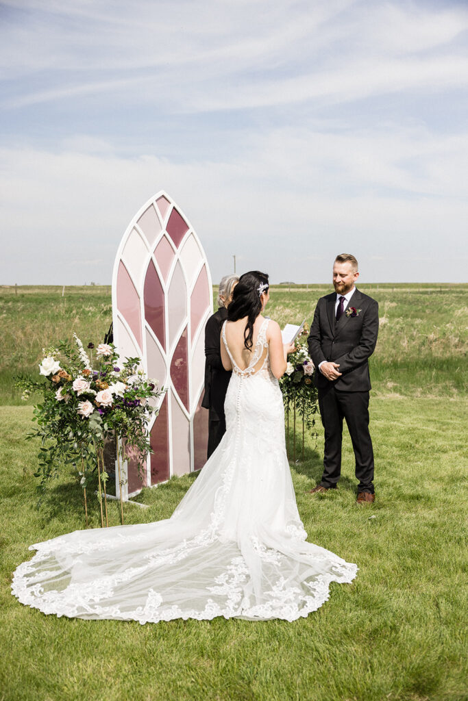A couple stands facing each other during a wedding ceremony outdoors, with a decorative arched doorway and floral arrangements. A Garden Wedding with Pops of Plum