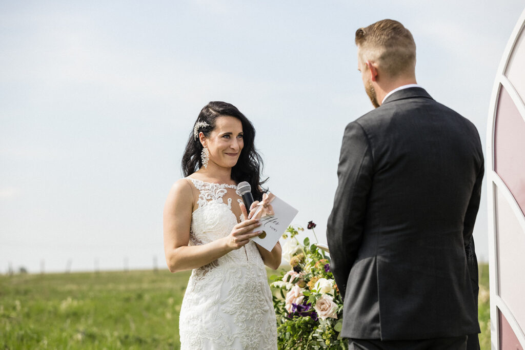 A bride speaking into a microphone and holding a note, facing the groom during an outdoor wedding ceremony.