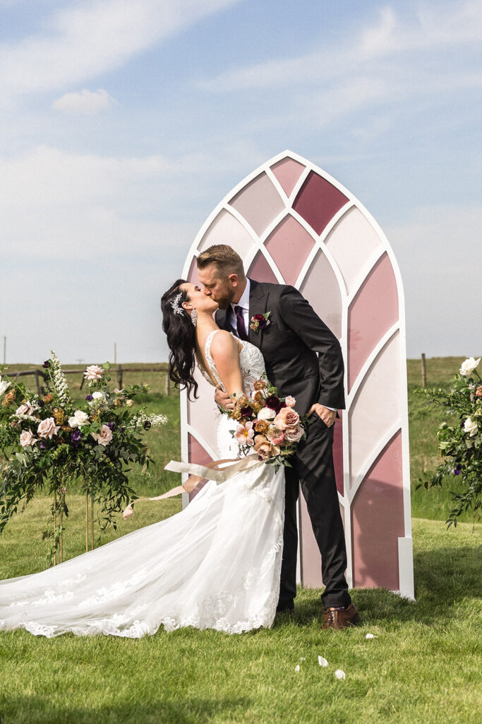 Bride and groom sharing a kiss beside a decorative arch at an outdoor wedding ceremony.