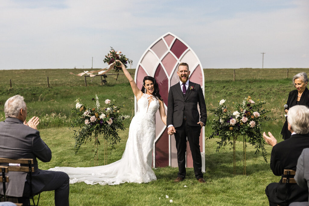 Bride and groom smiling and holding hands at outdoor wedding ceremony, with guests applauding and an arched floral decoration in the background. A Garden Wedding with Pops of Plum