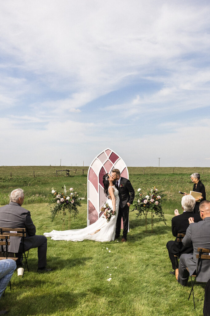 A bride and groom share a kiss at an outdoor wedding ceremony while guests look on.