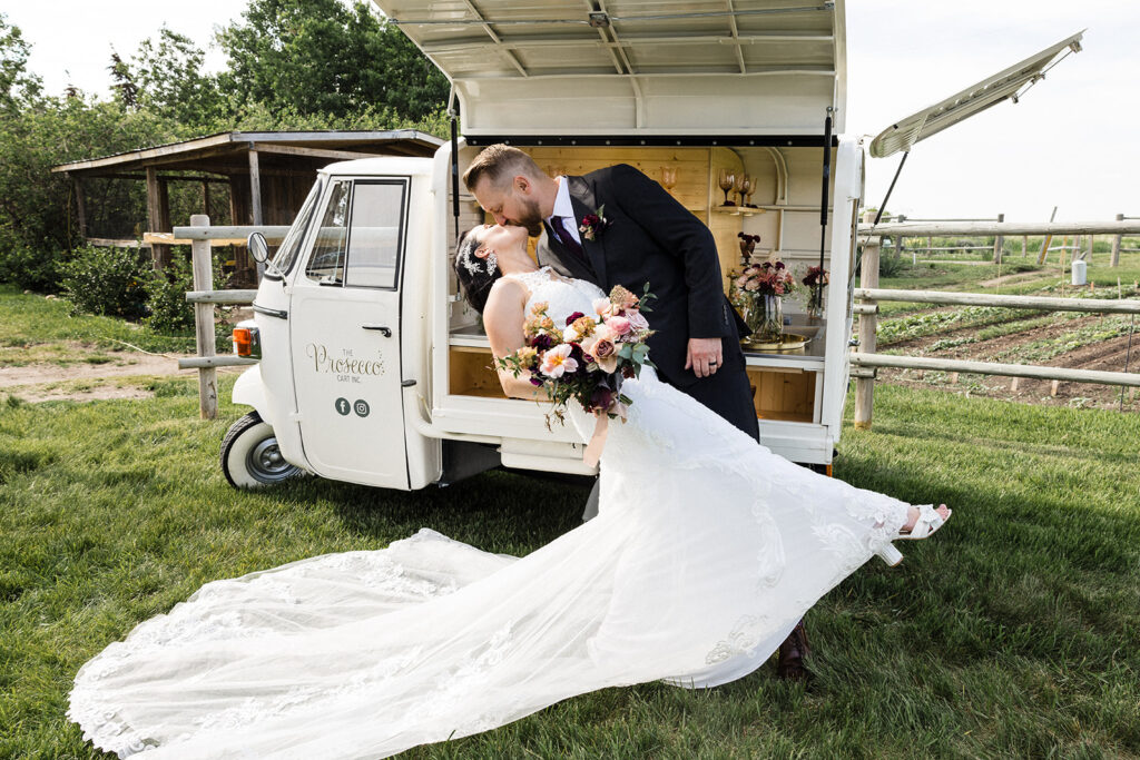 Bride and groom sharing a kiss beside a vintage van with an open side serving as a bar.