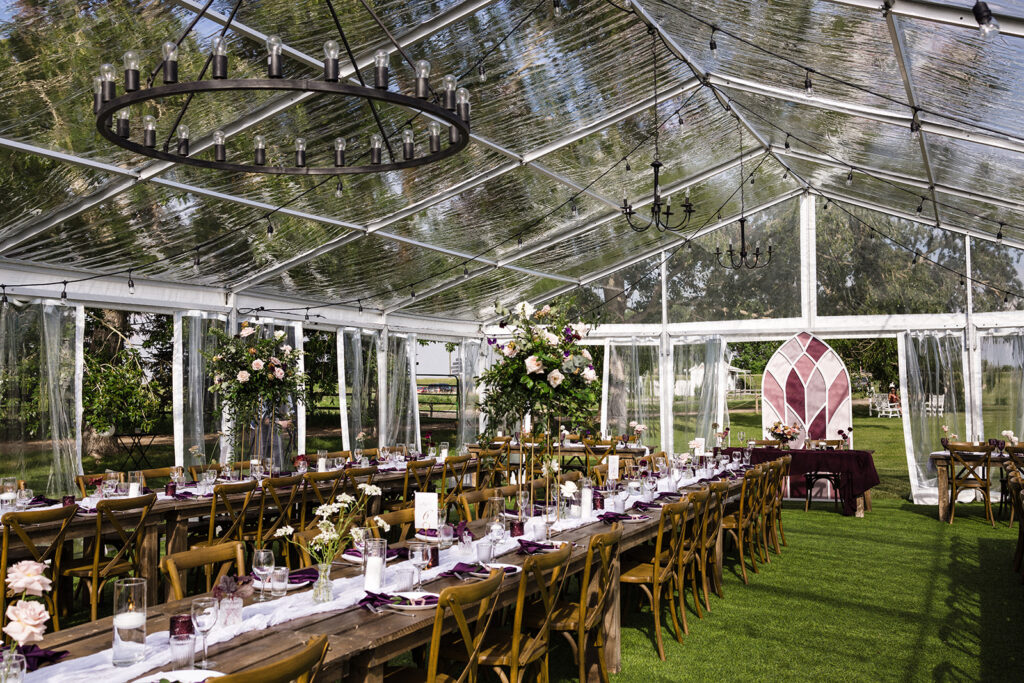Elegant outdoor event setup under a transparent tent with chandeliers, wooden tables, and floral arrangements.