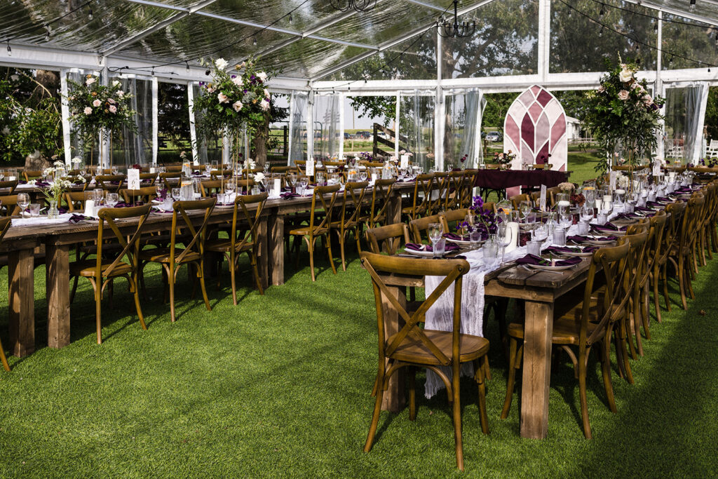 Elegant outdoor event setup with long wooden tables, floral centerpieces, and clear-top marquee.