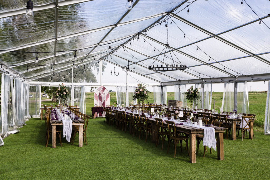 Elegant outdoor wedding setup under a clear marquee with white drapes, wooden tables, and floral decorations.