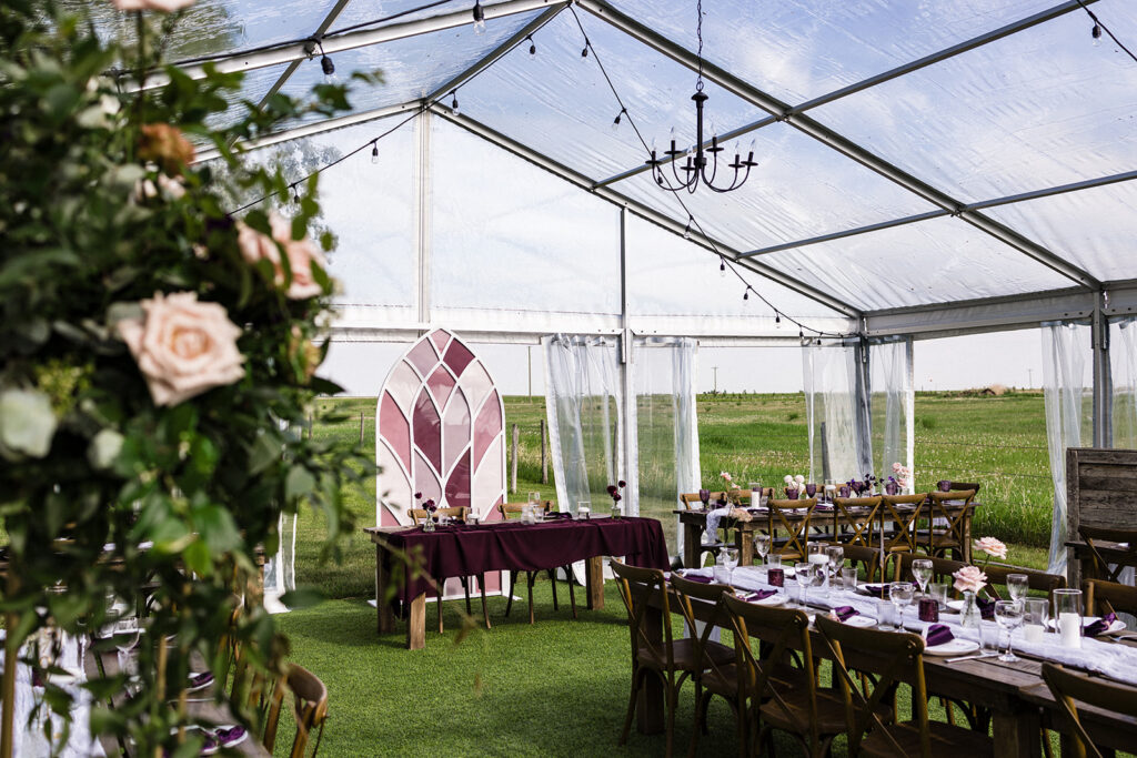 Elegant outdoor wedding reception setup under a clear tent with chandeliers, surrounded by natural scenery. A Garden Wedding with Pops of Plum