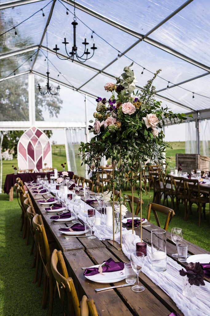 Elegant outdoor dining setup under a clear tent with floral centerpieces and wooden tables. A Garden Wedding with Pops of Plum