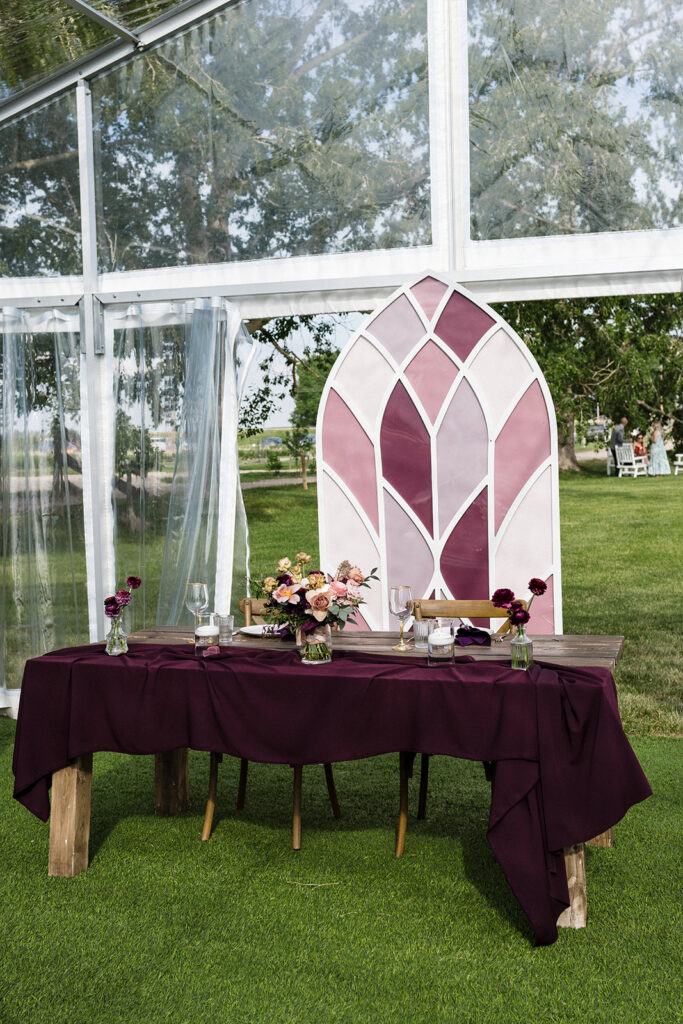 Elegant outdoor wedding table setting featuring a plum tablecloth, floral arrangements, and a stained glass backdrop. A Garden Wedding with Pops of Plum