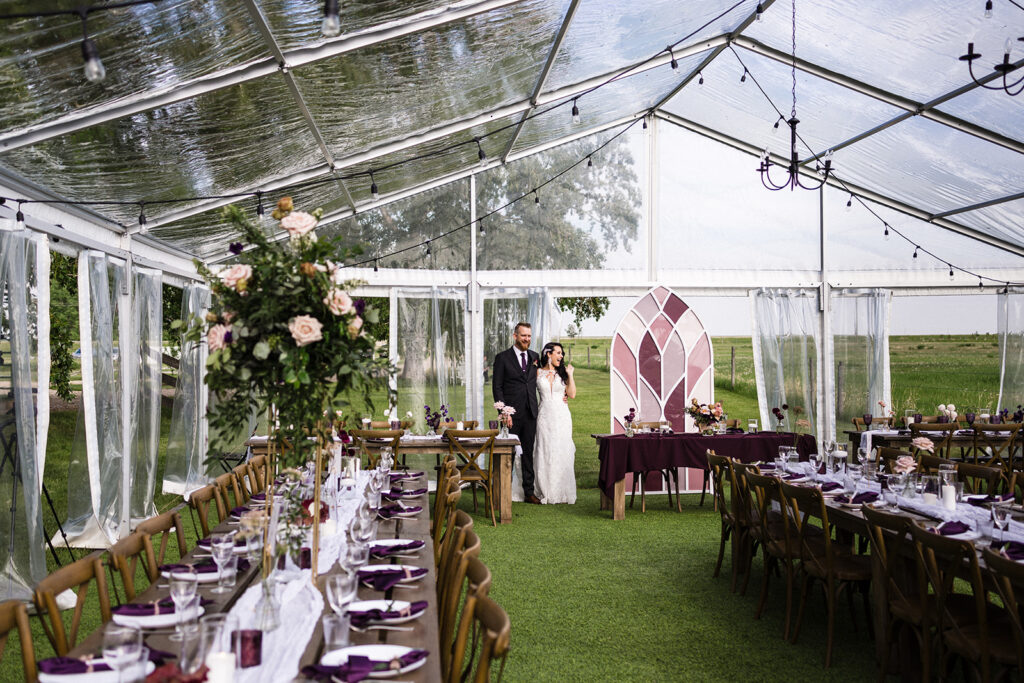 A bride and groom standing in a decorated clear tent set up for a wedding reception. A Garden Wedding with Pops of Plum