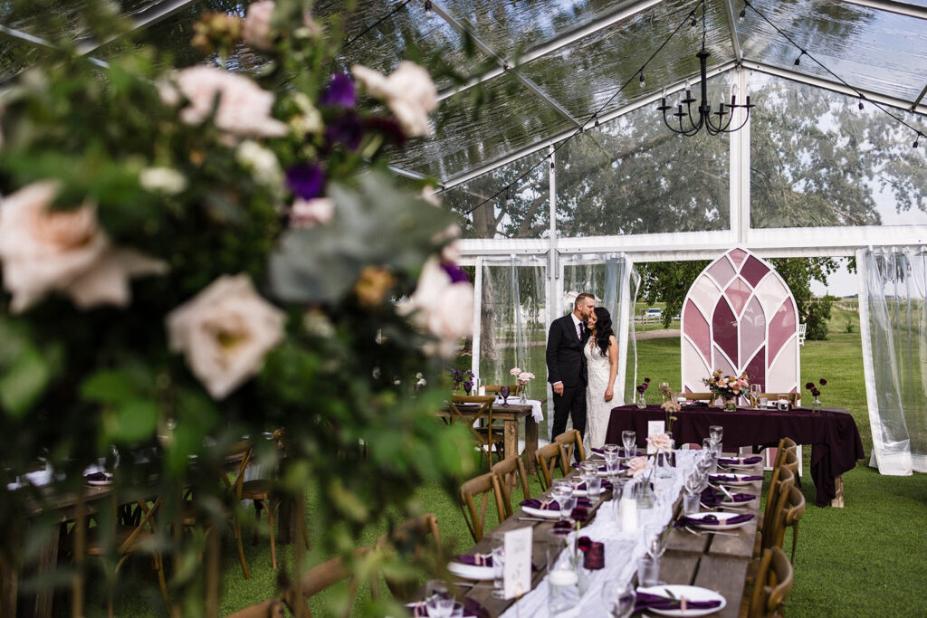 A couple embracing at the end of a decorated dining table set for an event inside a tent with a transparent roof. A Garden Wedding with Pops of Plum