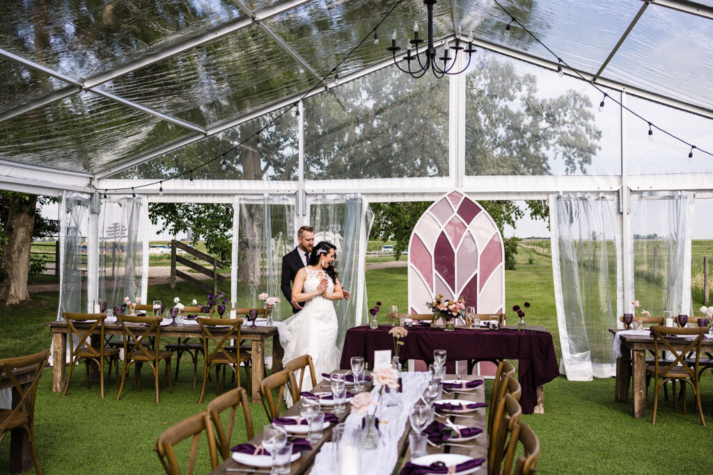 Couple standing together in a decorated wedding tent with set tables ready for guests. A Garden Wedding with Pops of Plum