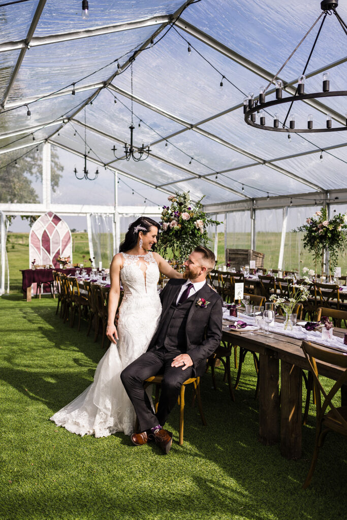 A couple dressed in wedding attire sharing a moment under a transparent tent at a decorated venue.