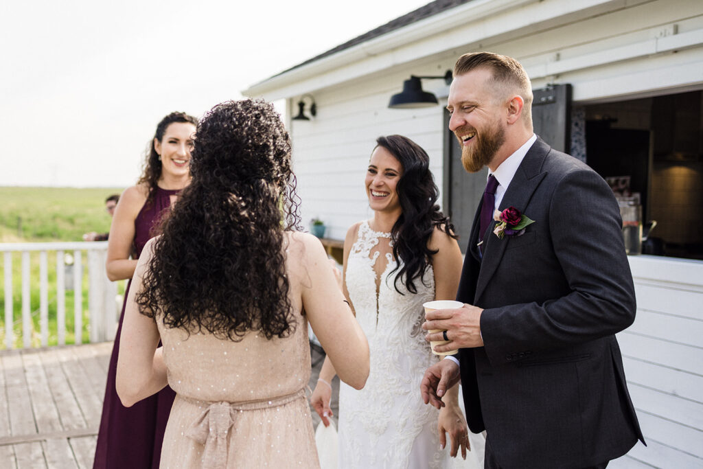 A joyful wedding group sharing a laugh outdoors, with a bride and groom in focus, holding drinks.
