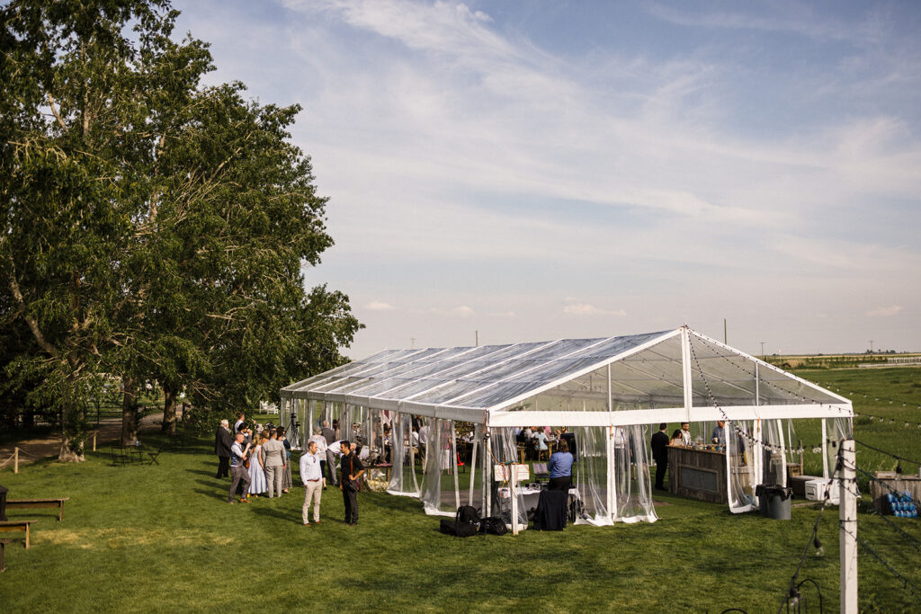 Outdoor event under a transparent tent with guests mingling on a green lawn.
