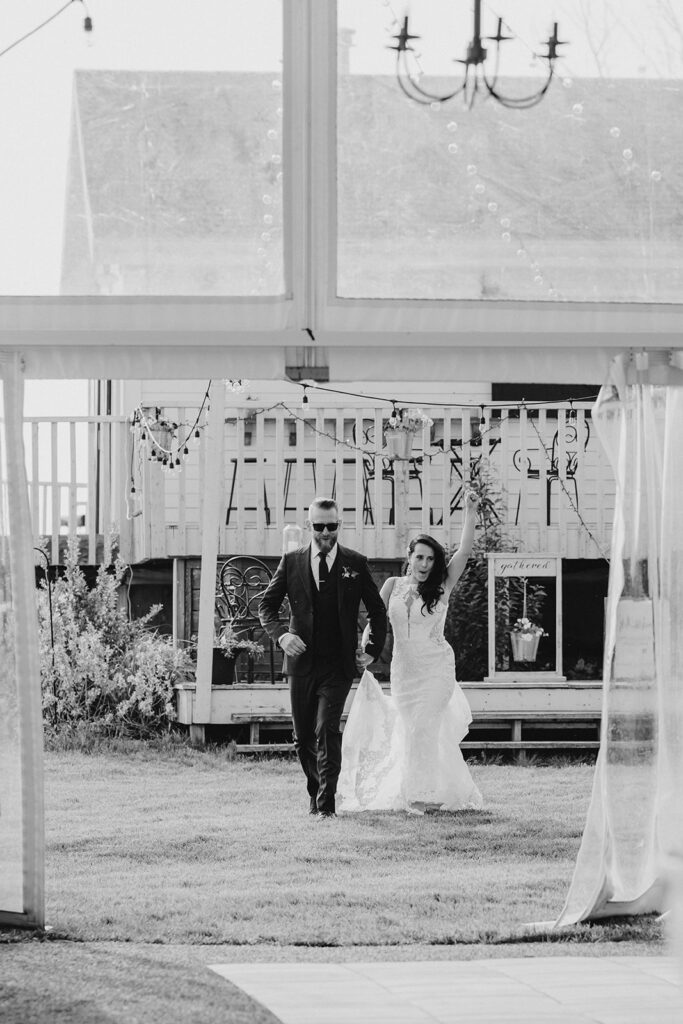 Bride and groom holding hands and walking confidently on a grassy outdoor area, with a decorative backdrop and white drapes in the foreground.