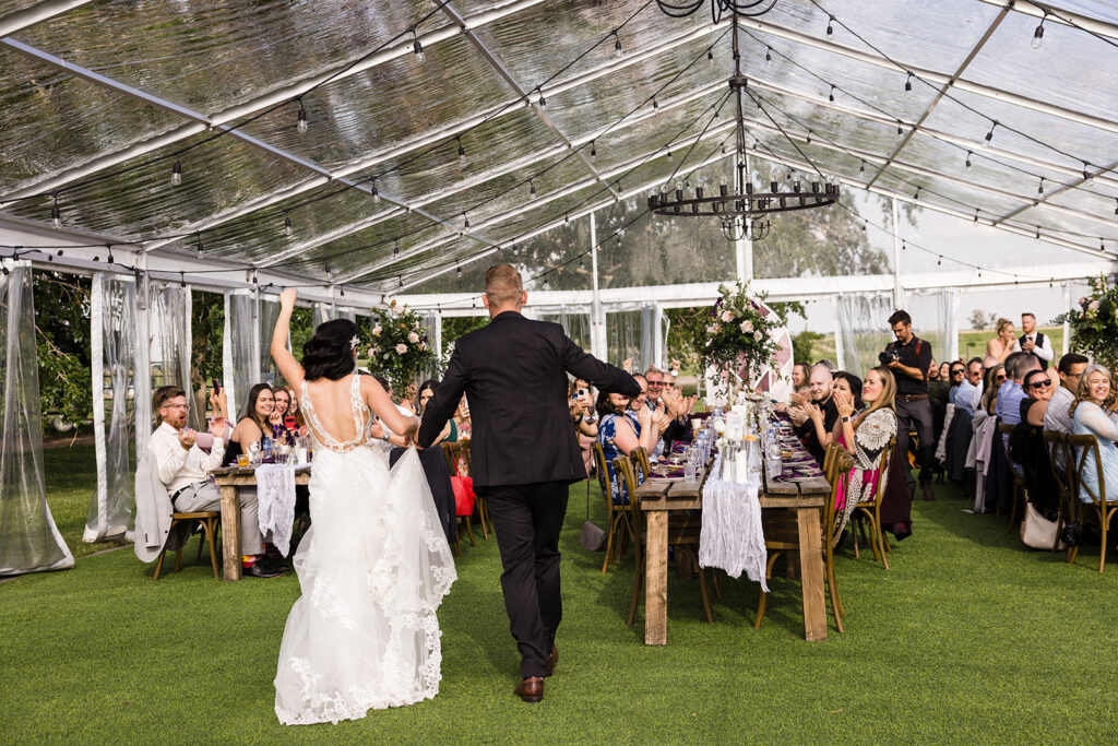 A couple walking down the aisle at their wedding reception under a transparent tent, with guests seated at tables on either side.