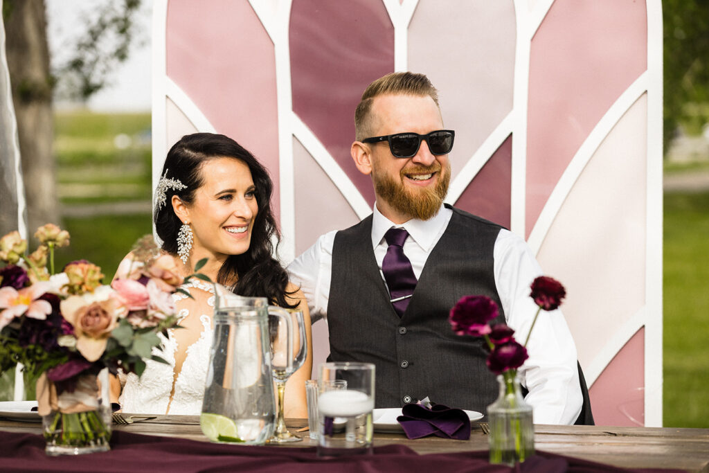 A smiling couple dressed in wedding attire seated at a decorated table outdoors.