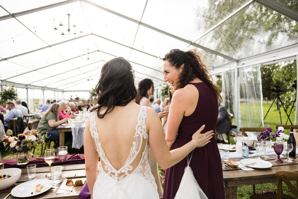Two women, one in a wedding dress, smiling and talking at an outdoor reception under a tent.