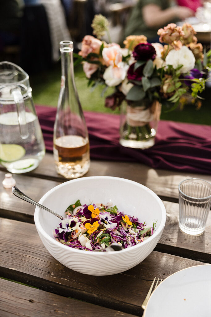 Elegant outdoor dining setting featuring a fresh salad garnished with edible flowers, with wine and floral centerpiece in the background.