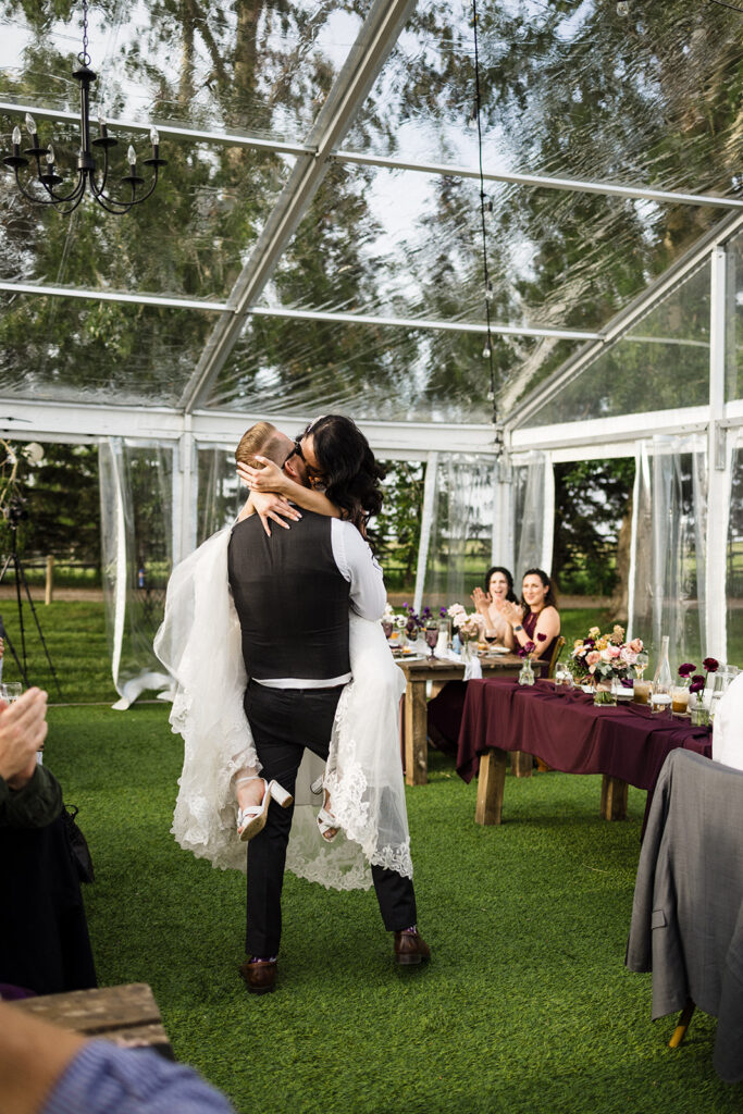 Bride and groom share a kiss in a transparent tent at an outdoor wedding reception while guests look on.