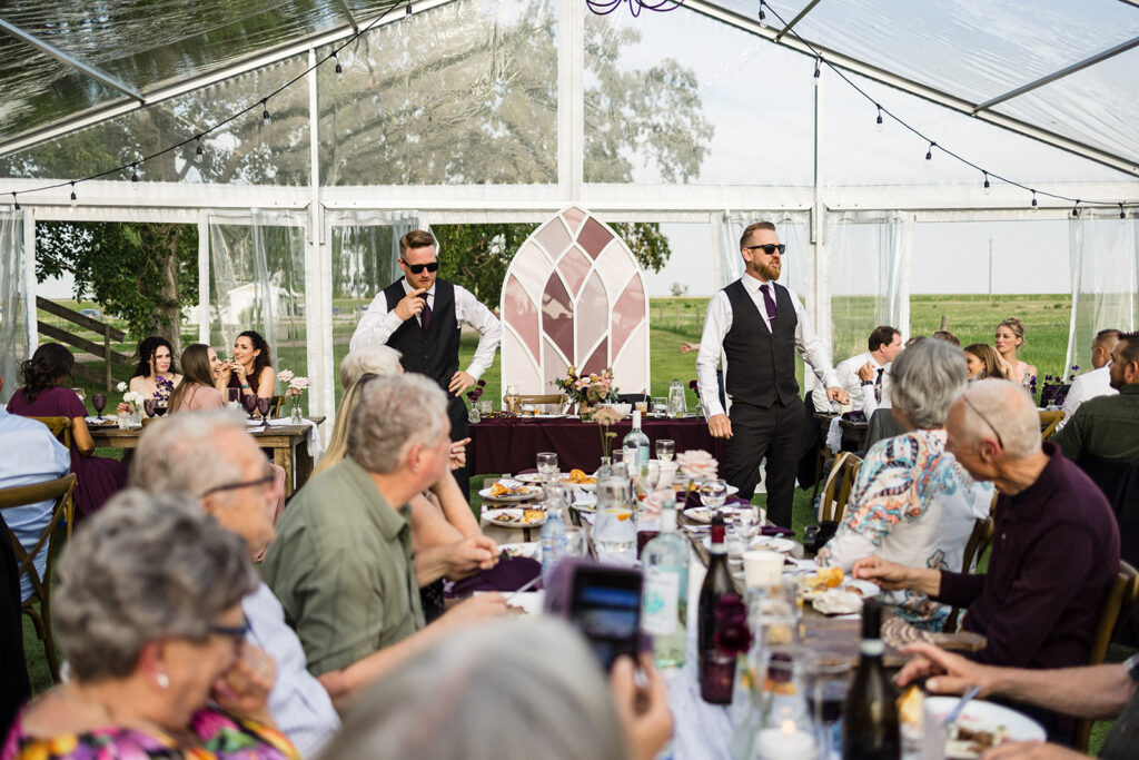 Two men standing and speaking at a wedding reception under a tent while guests are seated at tables.