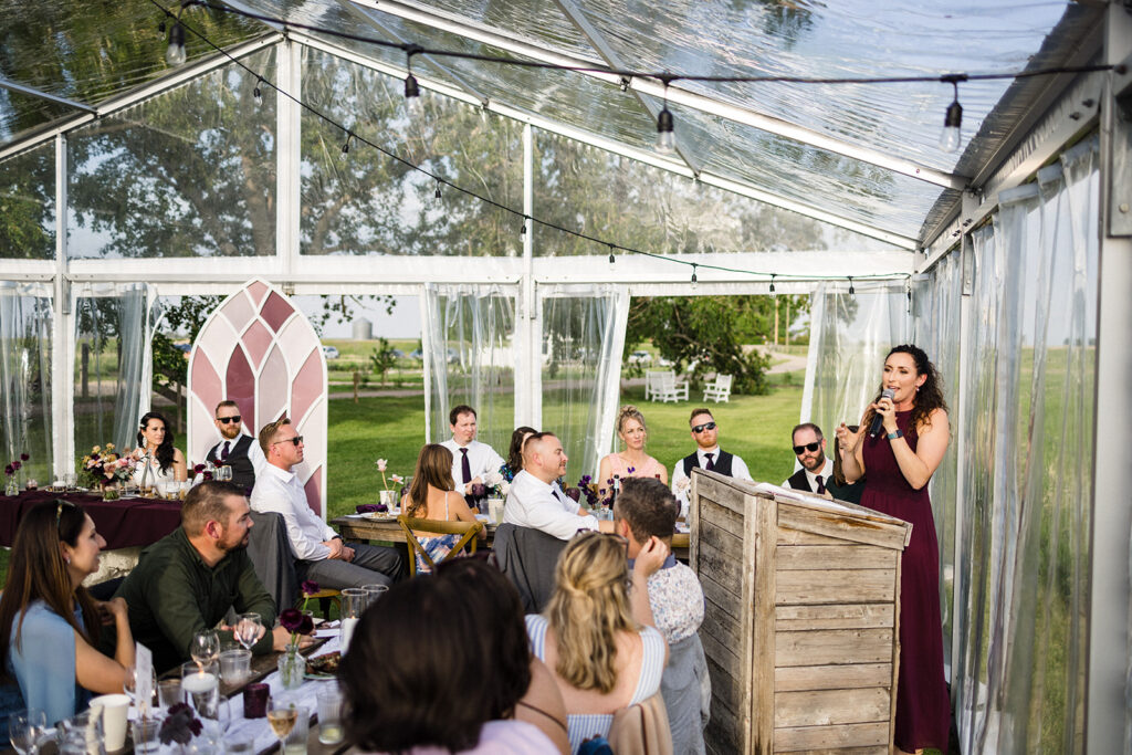 Woman giving a speech at an outdoor event under a clear tent with guests seated at tables.