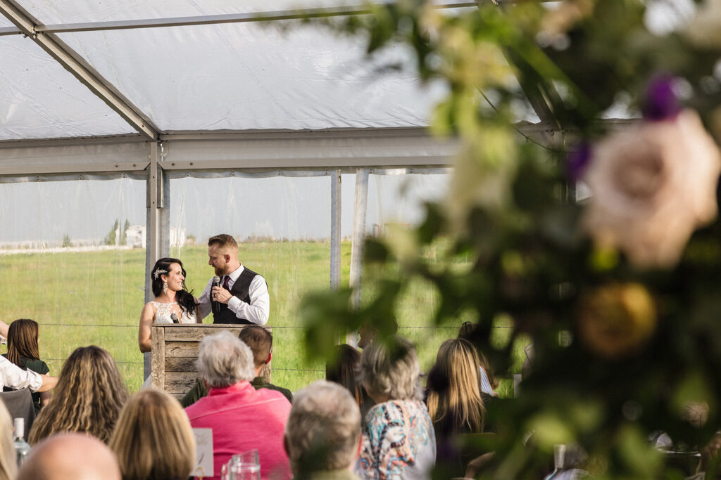A bride and groom exchanging vows in a tent at an outdoor wedding ceremony, as seen from behind the guests.