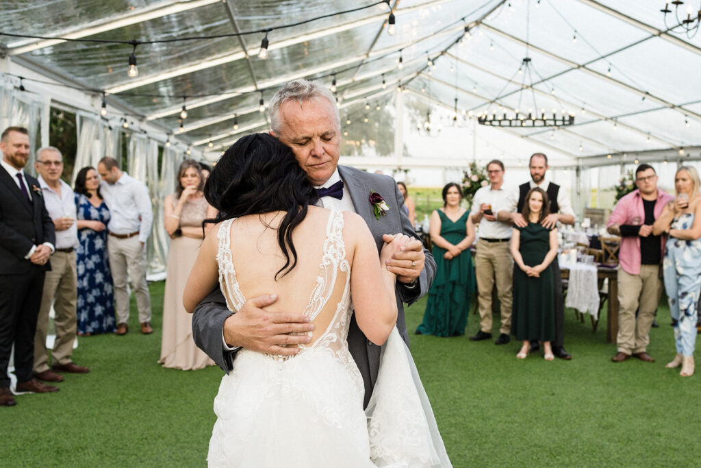 A father and daughter sharing a heartfelt dance at a wedding reception inside a greenhouse, with guests watching on.