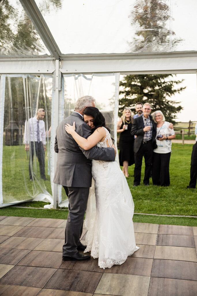 A bride and an older man sharing an emotional embrace on a dance floor at an outdoor wedding, with guests looking on.