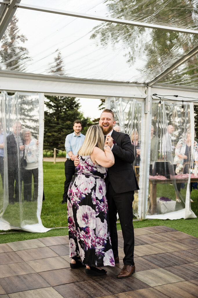 A couple dancing and laughing inside a clear tent with guests watching.