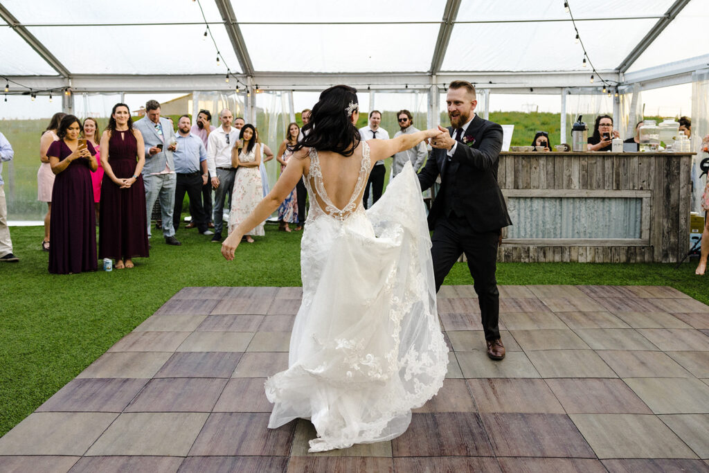 A bride and groom share a joyous dance on a wooden floor at their outdoor wedding reception while guests watch on.