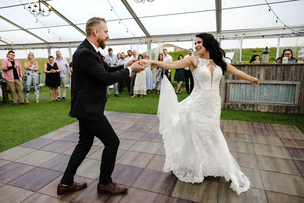 A bride and groom dancing joyfully on a wooden floor at their wedding reception, with guests looking on.