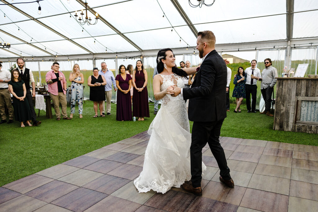 Bride and groom sharing a dance at an outdoor wedding reception under a tent while guests look on.